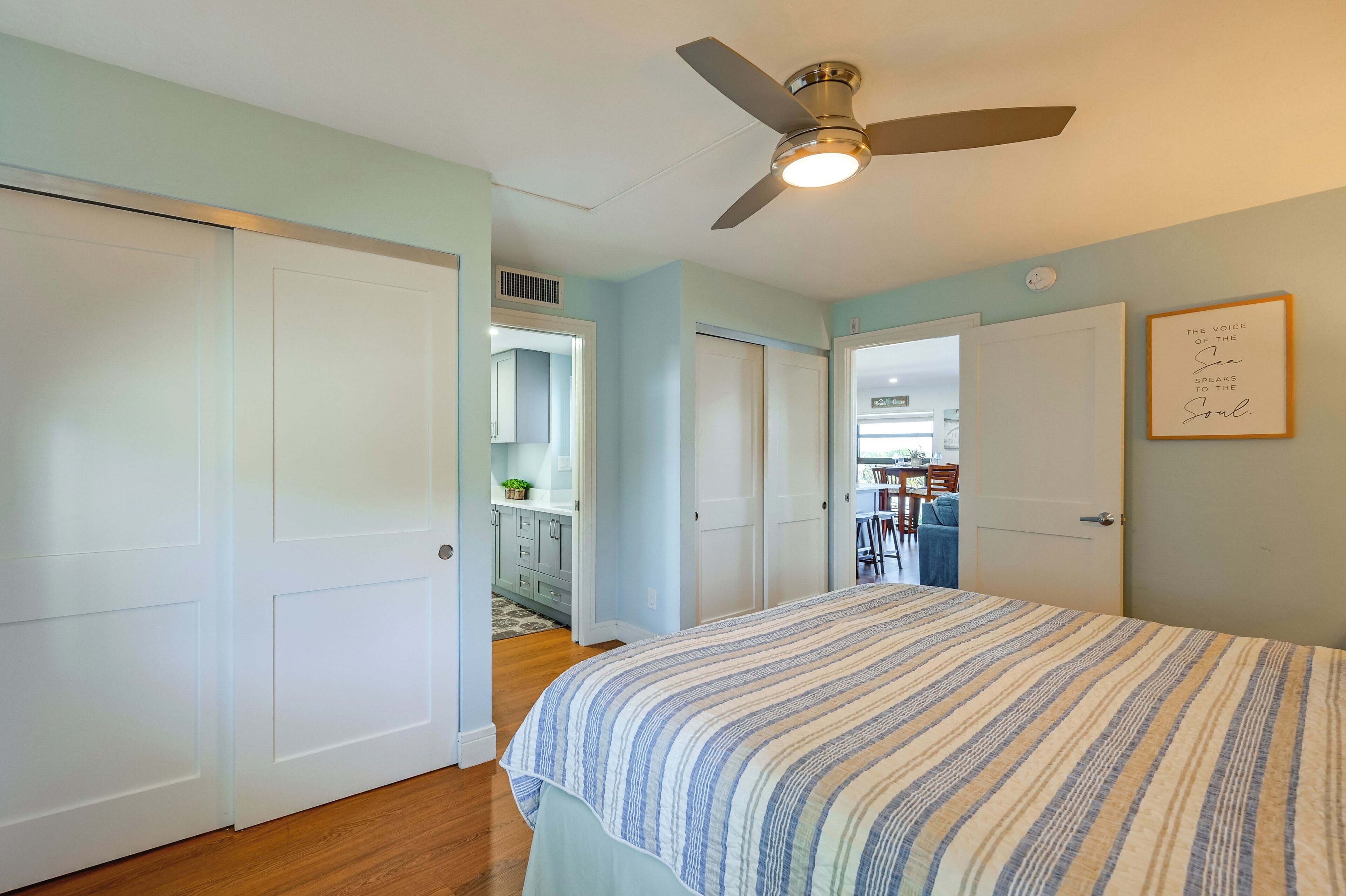 A bedroom with a bed, a ceiling fan, and a door leading to a kitchen area.