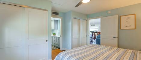 A bedroom with a bed, a ceiling fan, and a door leading to a kitchen area.