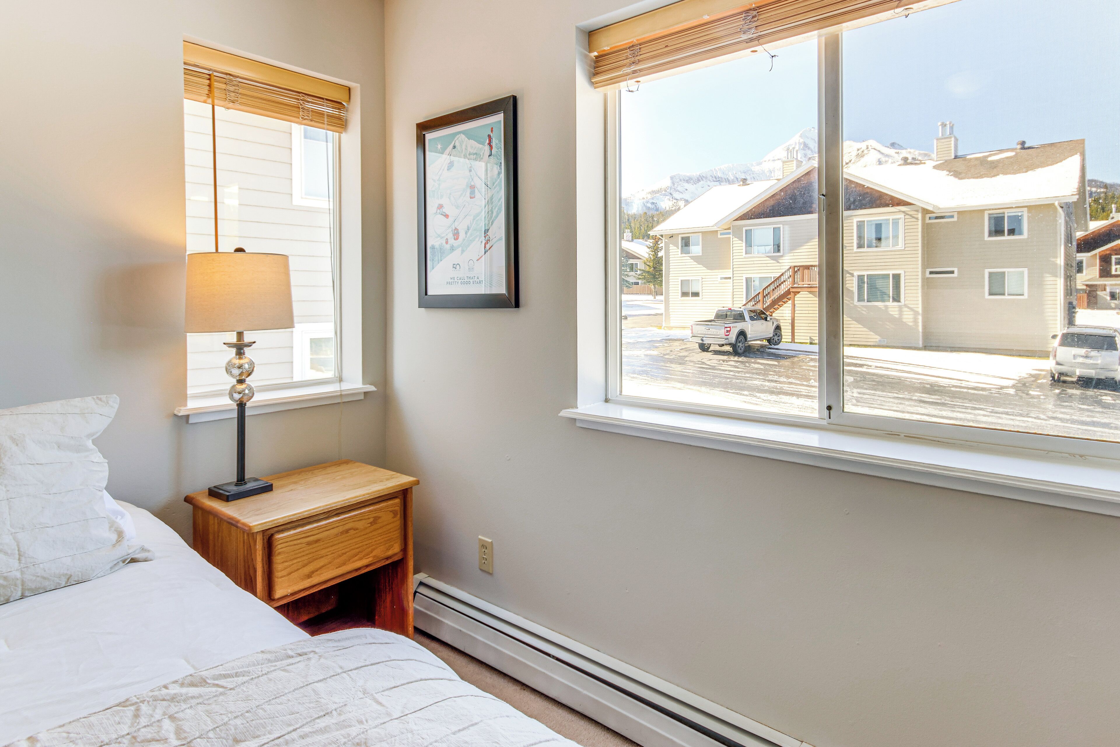 A bedroom with a bed, a nightstand, a lamp, a window with a view of a snowy mountain and buildings.