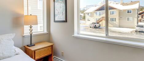 A bedroom with a bed, a nightstand, a lamp, a window with a view of a snowy mountain and buildings.