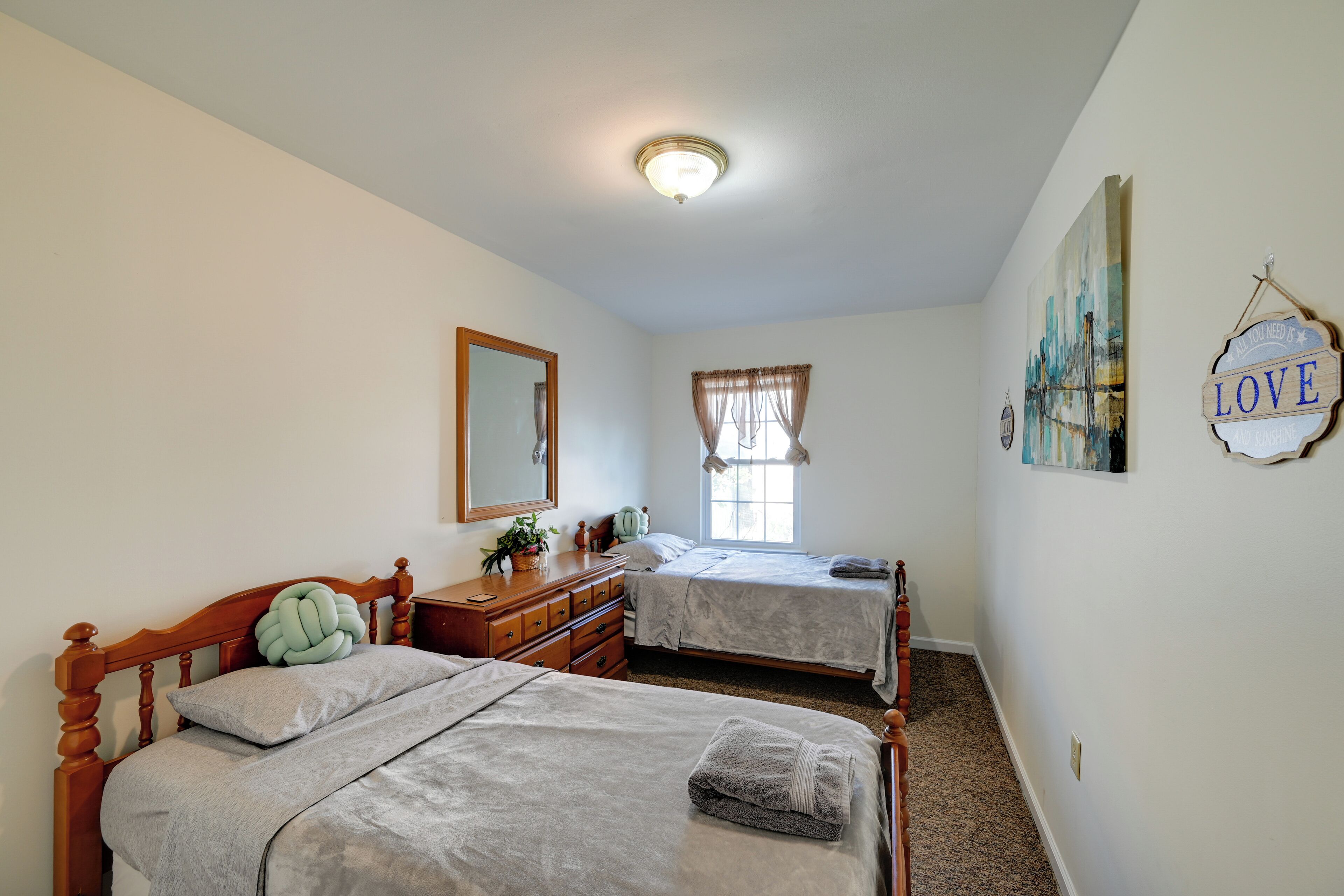 Une chambre avec deux lits simples, une commode, un miroir, une fenêtre avec des rideaux et un mur orné d’un panneau indiquant le mot « AMOUR ».