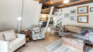 Ein geräumiger Wohnbereich mit einem weißen Sofa, einem grauen Sofa mit Felldecke und einem gemusterten Sessel. Außerdem sind eine Holzkommode und eine Lampe zu sehen.