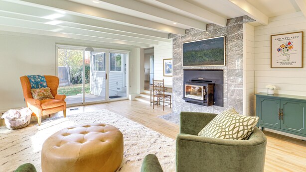 Amplia sala de estar con chimenea, un sillón verde, un reposapiés color beige y un ventanal con vistas al exterior.