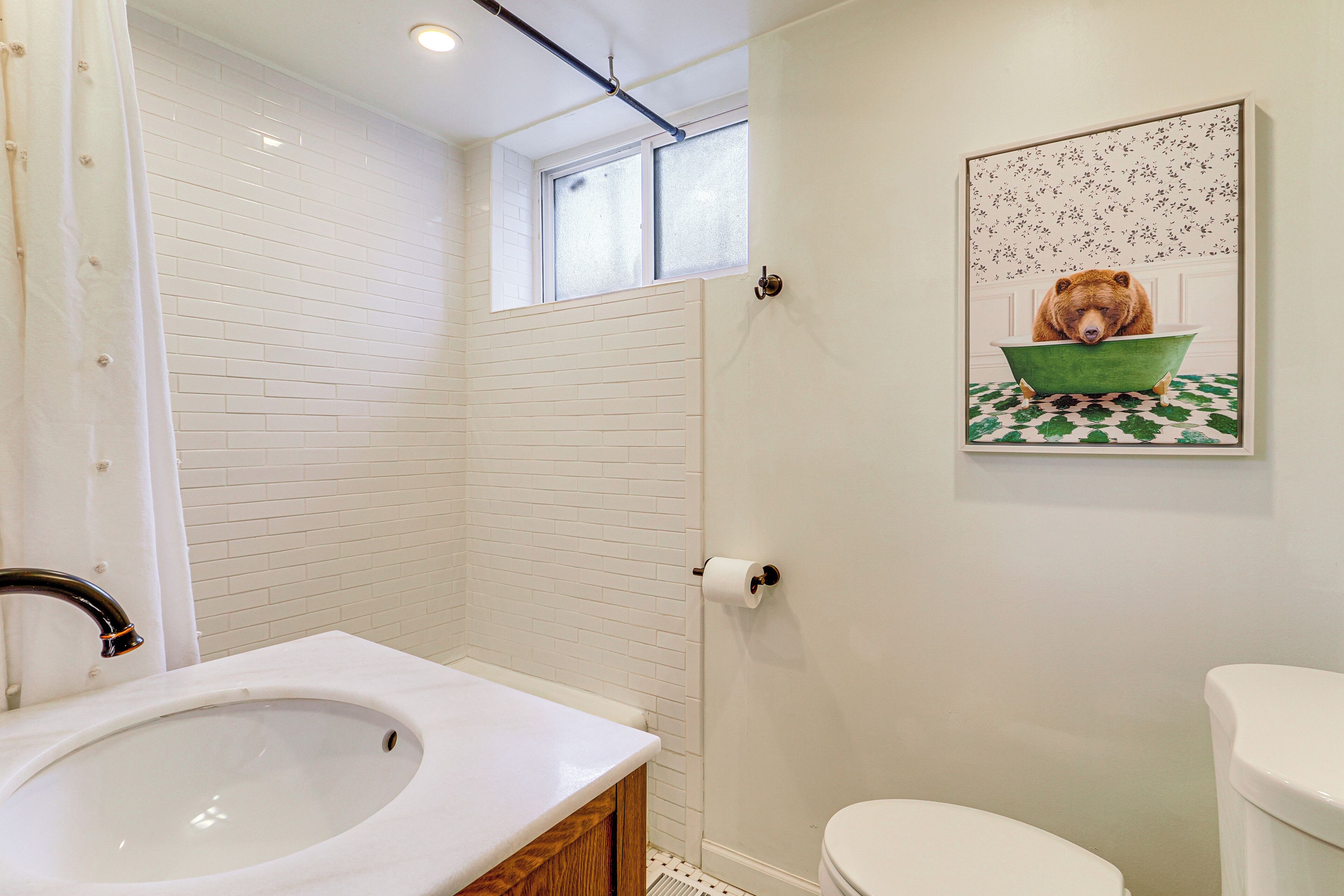 Ein Badezimmer mit Waschbecken, Toilette und Dusche mit Vorhang. Über der Dusche befindet sich ein Fenster und an der Wand hängt ein Bärgemälde.