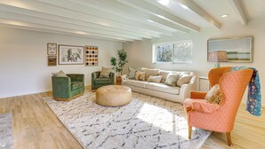 Un salón amplio con techo alto, un ventanal grande y diversas opciones de asiento, incluyendo un sillón verde, un sofá color beige y un sillón naranja.