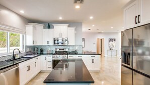 A modern kitchen with white cabinets, a central island, stainless steel appliances, and a large window providing natural light. - Riverfront Fort Myers Luxury Home w/ Heated Pool! (Fort Myers)
