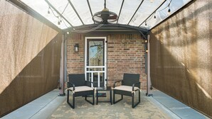 Een overdekt terras met twee stoelen en een tafel, een bakstenen muur en een deur met ruit.