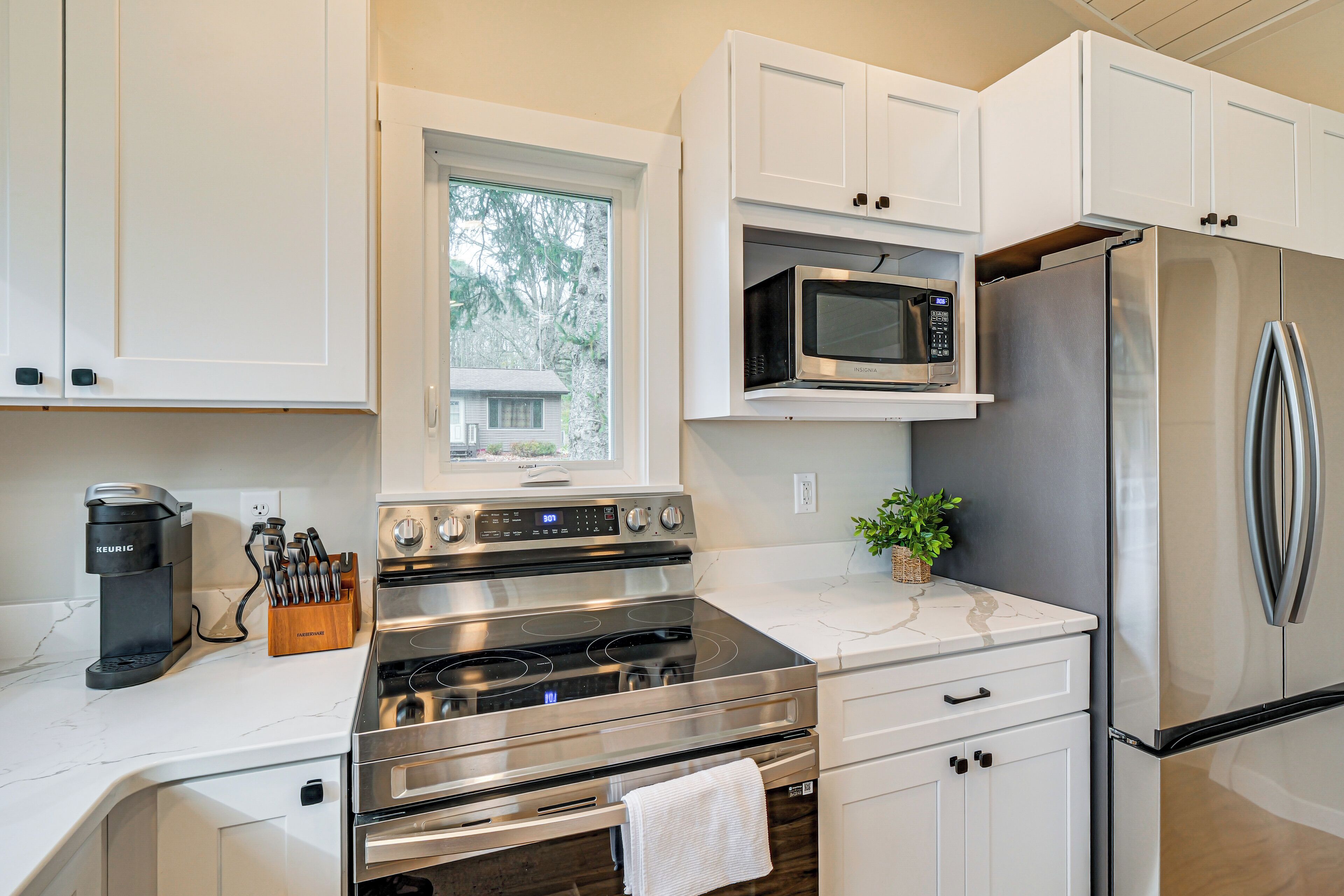 A modern kitchen with white cabinets, a stainless steel refrigerator, a microwave, a Keurig coffee maker, and a window with a view of trees.