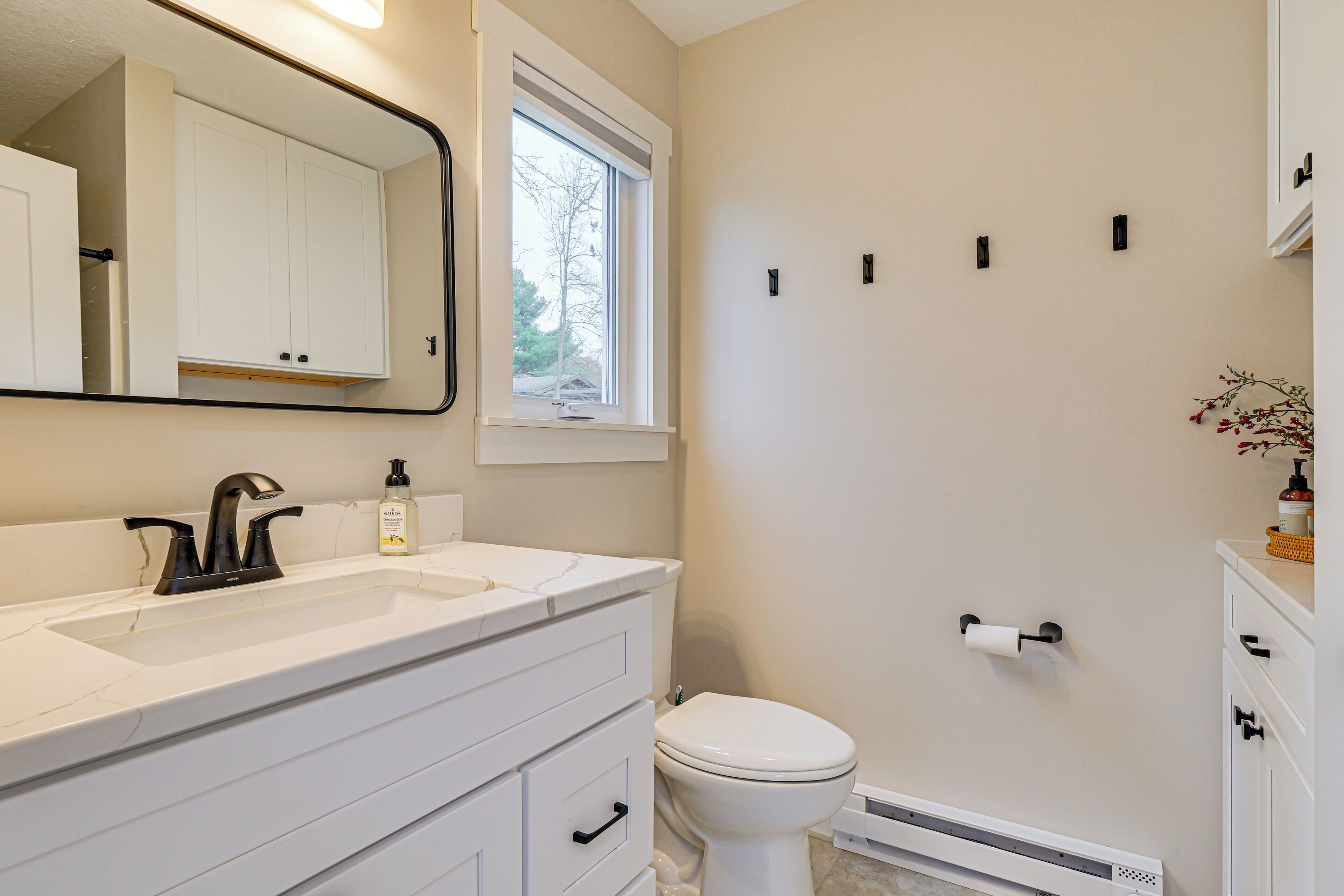 A bathroom with a white sink, black faucet, white toilet, and a window above the toilet. There are three black wall-mounted lights and a white cabinet with drawers.