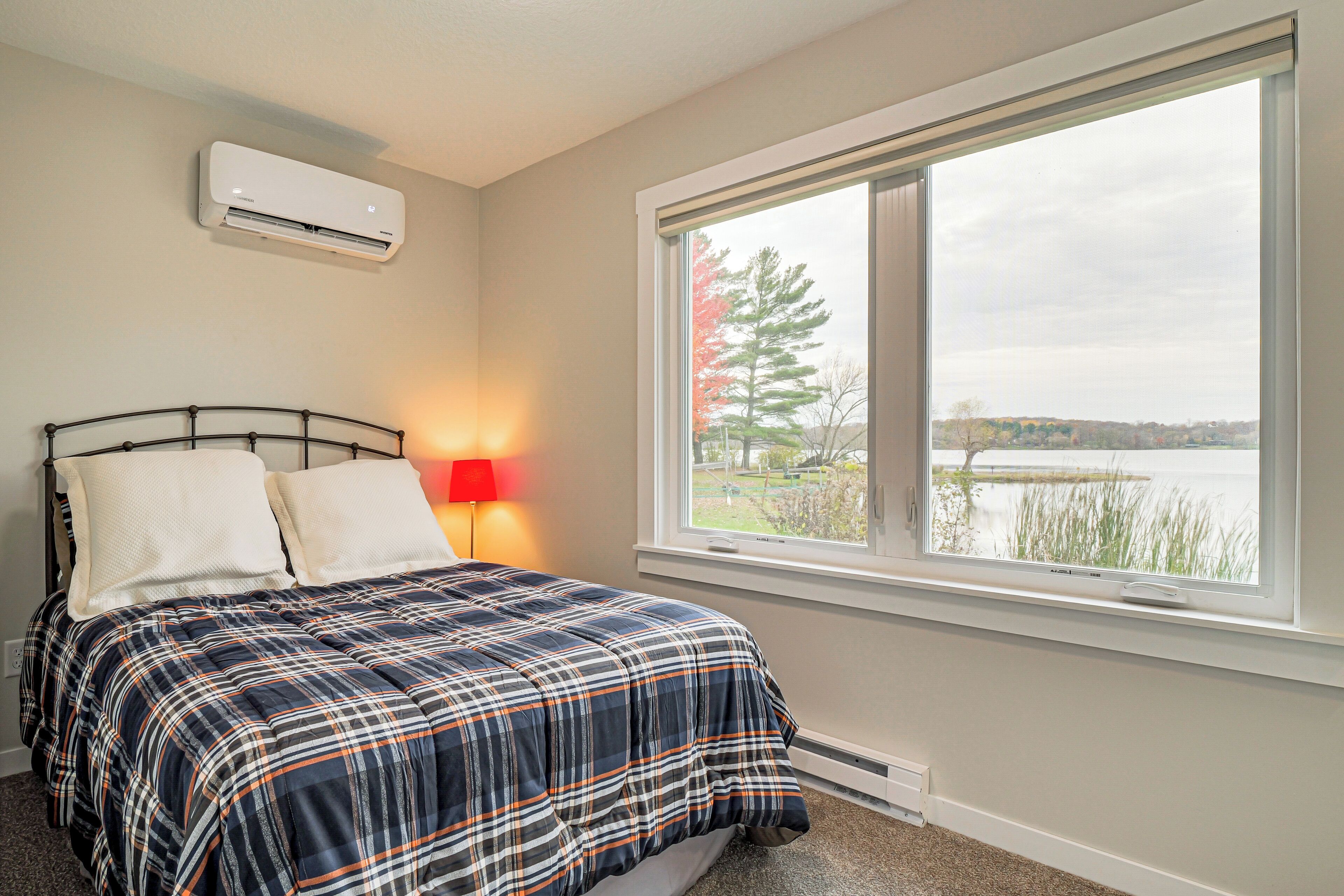A bedroom with a bed, a window with a view of a lake, an air conditioner, and a red lamp.