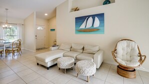 A living room with a sofa, two ottomans, a wicker chair, a painting of sailboats, and a dining area with chairs and a table. - Family Haven w/ Pool! 8 Mi to Warm Mineral Springs (North Port)