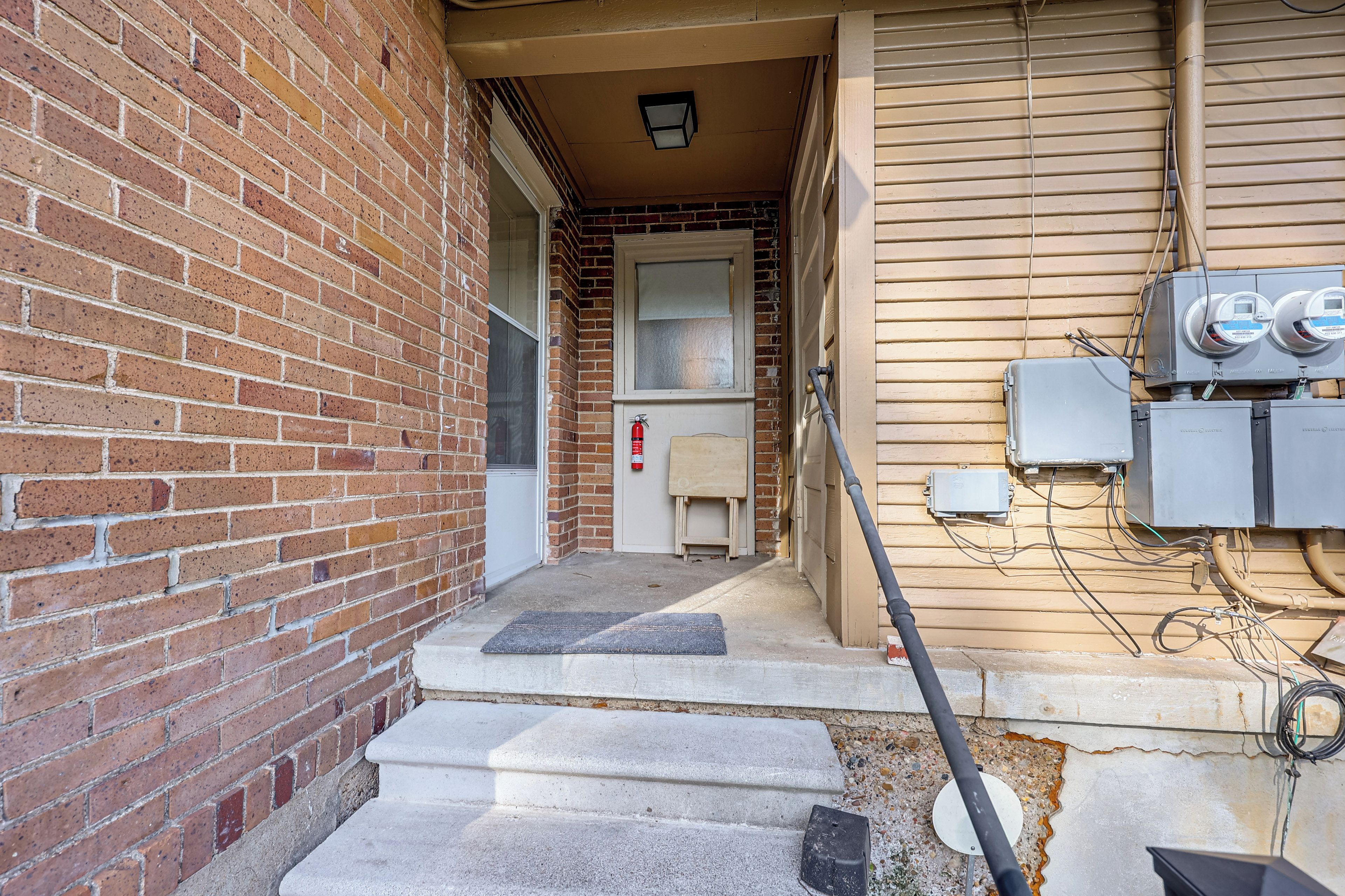 A narrow alleyway with a brick wall on one side and a beige building on the other. There is a fire extinguisher mounted on the wall, a door with a window, and electrical boxes attached to the building.
