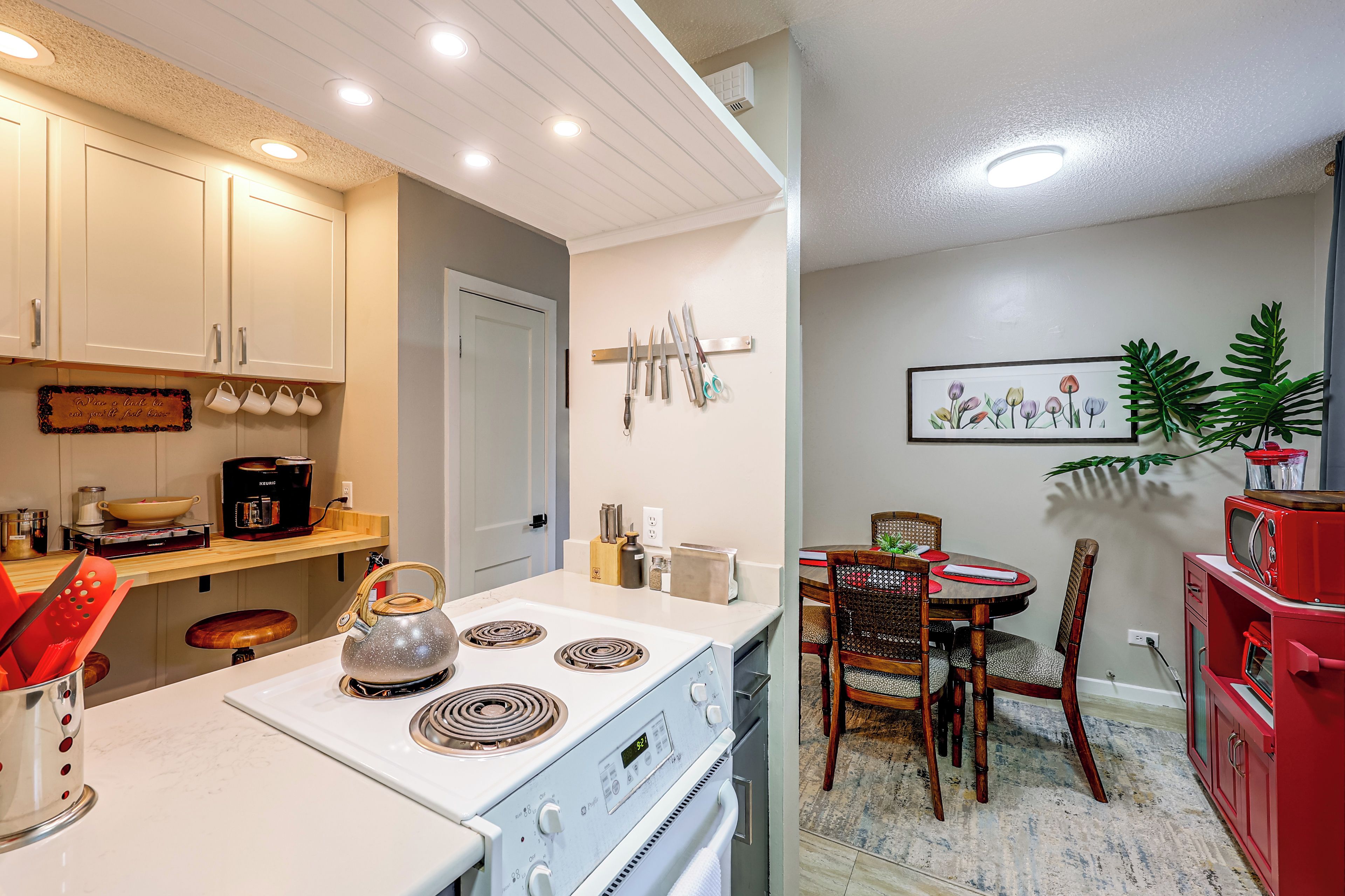 A modern kitchen with white cabinets, a stove, and a dining area with a red cabinet and a framed picture on the wall.