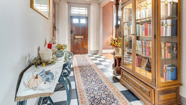 A hallway with a checkered floor, a table with a tablecloth, a wooden cabinet with glass doors, and a door in the background. - Pet-friendly Historic Library Home on Route 66 (Virden)