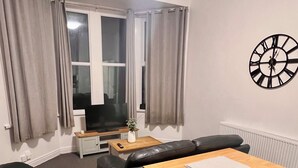 Un soggiorno con un divano, un tavolo in legno, una TV e una sveglia appesa al muro.
