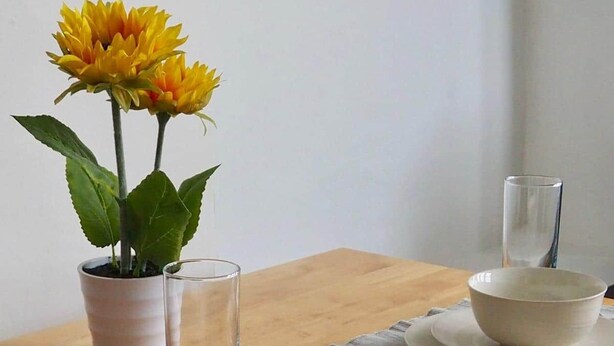 Un tavolo da pranzo apparecchiato con ciotole, piatti e posate bianche, un bicchiere e un vaso con fiori gialli.