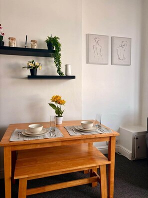 A dining area with a wooden table set for two, a potted plant, and a wall-mounted shelf with decorative items. - Modern 2 bed flat in City Centre, High speed WiFi (Cardiff)