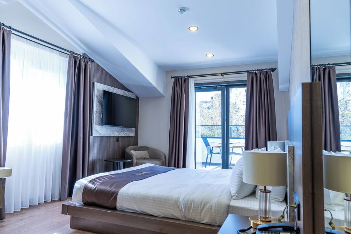 Camera d'albergo moderna con un grande letto, una TV a schermo piatto montata a parete, una scrivania con lampada e vista all'esterno attraverso ampie finestre.