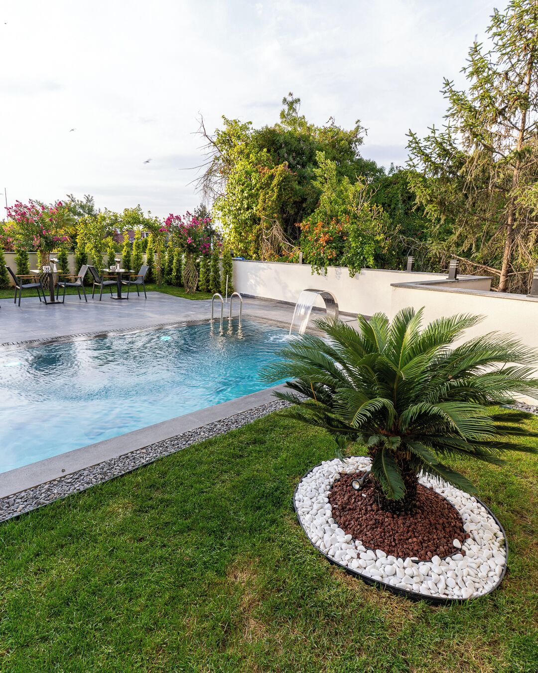 Una piscina con caída de agua, rodeada de un césped cuidado y una palmera.