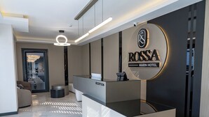 A modern reception desk with a sign for Rossa Marlin Hotel, a lounge area with seating, and a large suitcase on the floor. - ROSSA MARIN HOTEL (Darica)