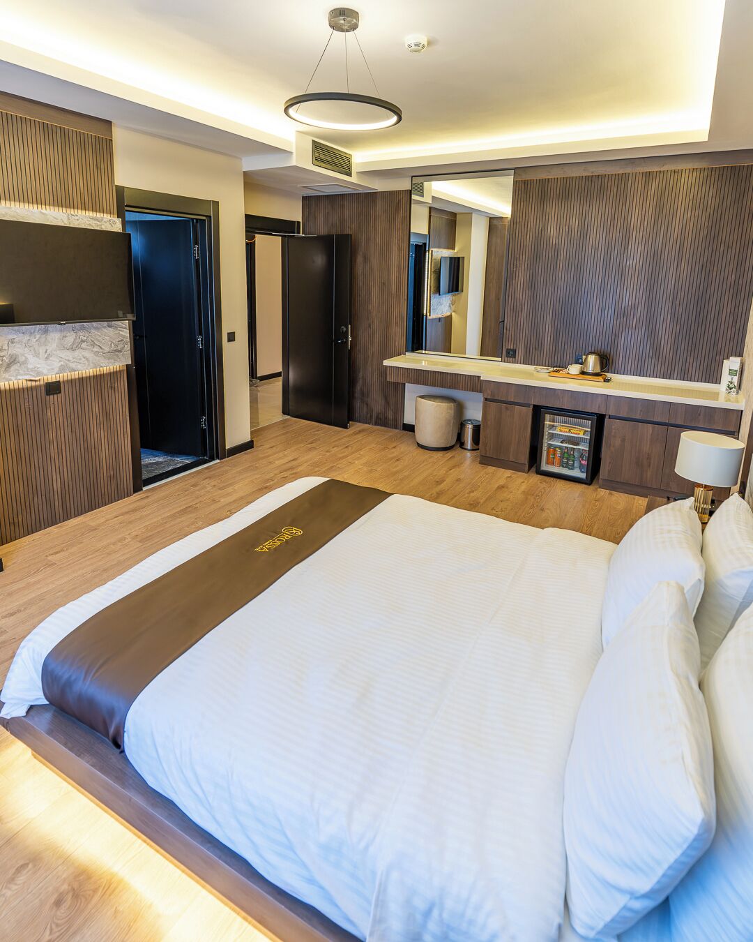 A modern hotel room with a large bed, a built-in desk, a minibar, and a large mirror.