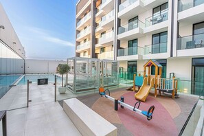 Ein Spielplatz auf dem Dach mit Rutsche und Wippe, umgeben von einem Glasgeländer und einem Gebäude mit Balkonen.