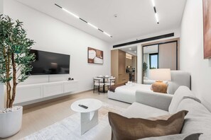 A modern hotel room with a large flat-screen TV, a sofa, a coffee table, a dining table, and a potted plant. - Stayshort - Studio Unit in JVC Sleeps 4 With Balcony (Dubai)