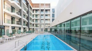 Ein moderner Innenpool mit klarem Wasser, umgeben von Liegestühlen und Sonnenschirmen, neben einem mehrstöckigen Gebäude mit Balkonen.