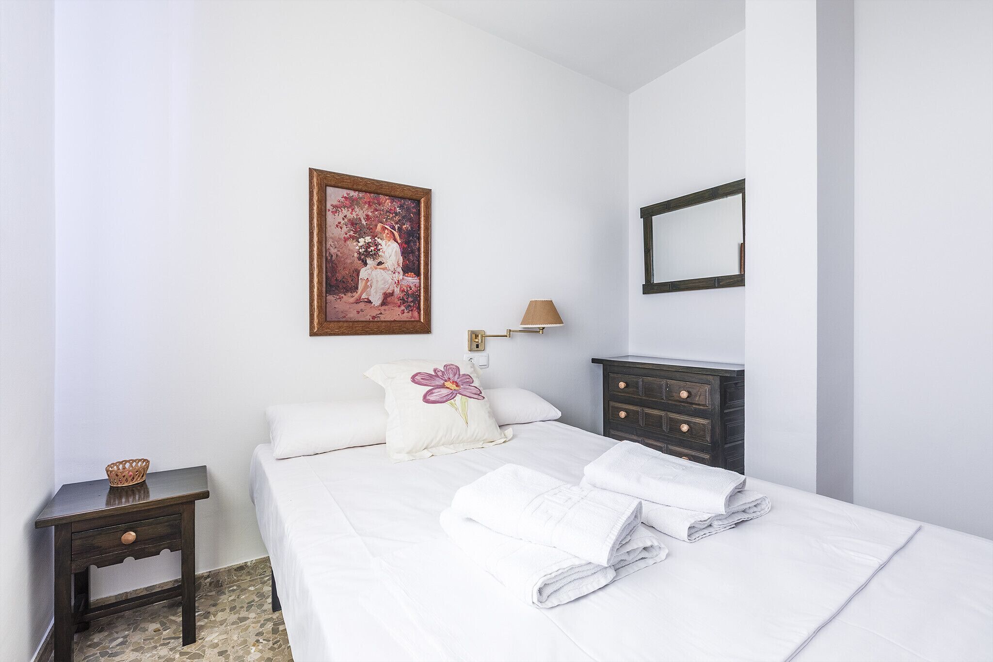 Una cama bien hecha con sábanas blancas y una toalla doblada, una mesita de noche con una cesta, un aparador de madera oscura, un cuadro en la pared y un espejo fijado a la pared.