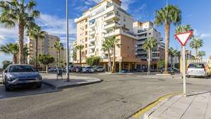 Un edificio de varios pisos con balcones, coches aparcados y palmeras.