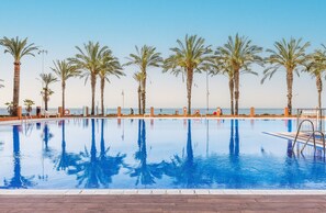 Ein Swimmingpool mit klarem, blauem Wasser, umgeben von Palmen und Blick aufs Meer.