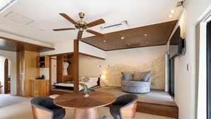 A modern living room with a round wooden table, two grey armchairs, a ceiling fan, a stone wall, and a grey sofa. - THE GARDEN Bise (Motobu)