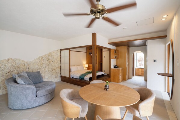 A modern hotel room with a stone accent wall, a grey sofa, a wooden dining table, and a ceiling fan. - THE GARDEN Bise (Motobu)