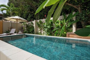 A rectangular swimming pool with clear blue water, surrounded by a tiled deck. There are lounge chairs and a white umbrella in the background. - THE GARDEN Bise (Motobu)