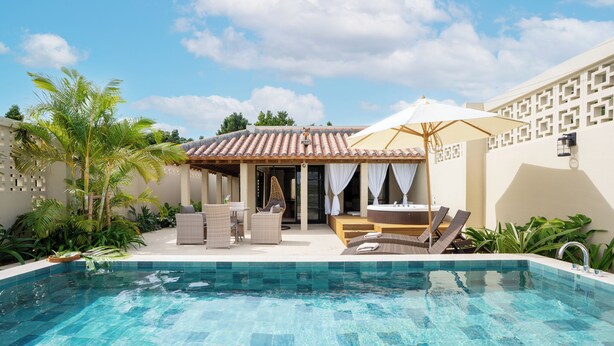 A swimming pool with tiled flooring, surrounded by lounge chairs and a covered seating area with an umbrella. - THE GARDEN Bise (Motobu)