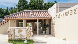 A building with a tiled roof, a stone wall with a sign reading "The Garden Cafe", and a paved entrance area with potted plants. - THE GARDEN Bise (Motobu)