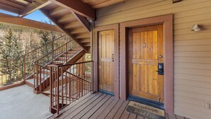 A wooden staircase leading to a balcony with a metal railing, a wooden door with a doormat, and a view of trees. - Walk to the Lifts! Cozy Condo with Access to Outdoor Hot Tubs (Keystone)
