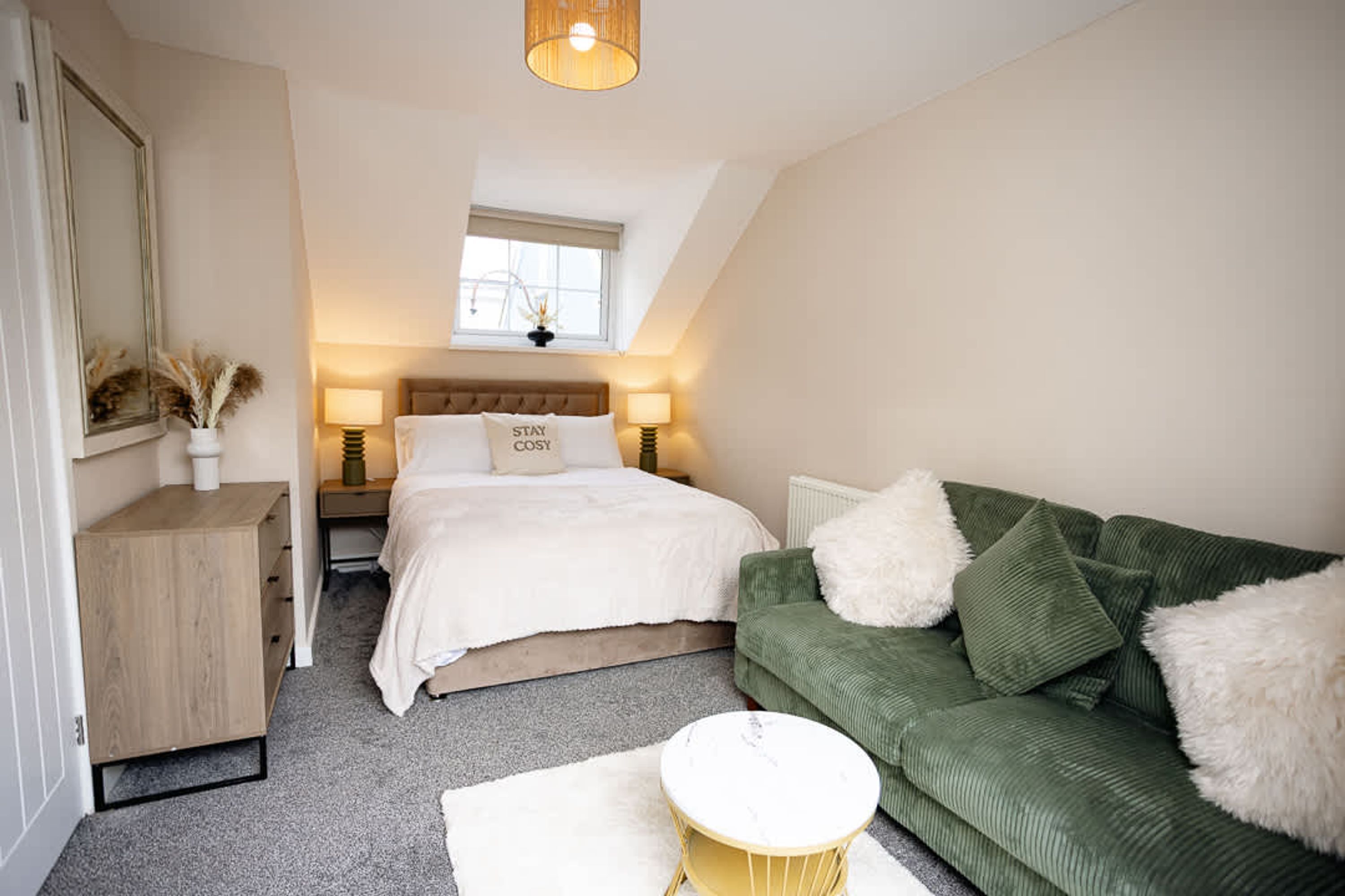 A bedroom with a bed, a green sofa, a small round table, a nightstand, a lamp, a window, and a mirror.
