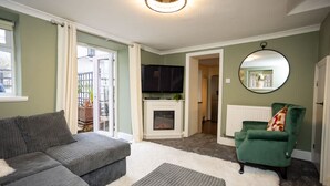 A living room with a grey sofa, a green armchair, a fireplace, and a television. - Rose Cottage - 2 Bedroom Cottage - Penally (Tenby)