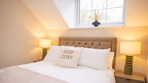 A neatly made bed with a pillow that reads "STAY COSY," two bedside lamps, a window with a view of trees, and a small nightstand with a lamp. - Rose Cottage - 2 Bedroom Cottage - Penally (Tenby)