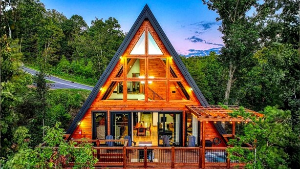 Una cabaña de madera con techo triangular, ventanas grandes y una terraza con mobiliario exterior.