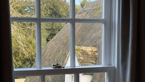Een raam met een hartvormige hendel, uitzicht op een huis met rieten dak, en een gedeeltelijk open gordijn.