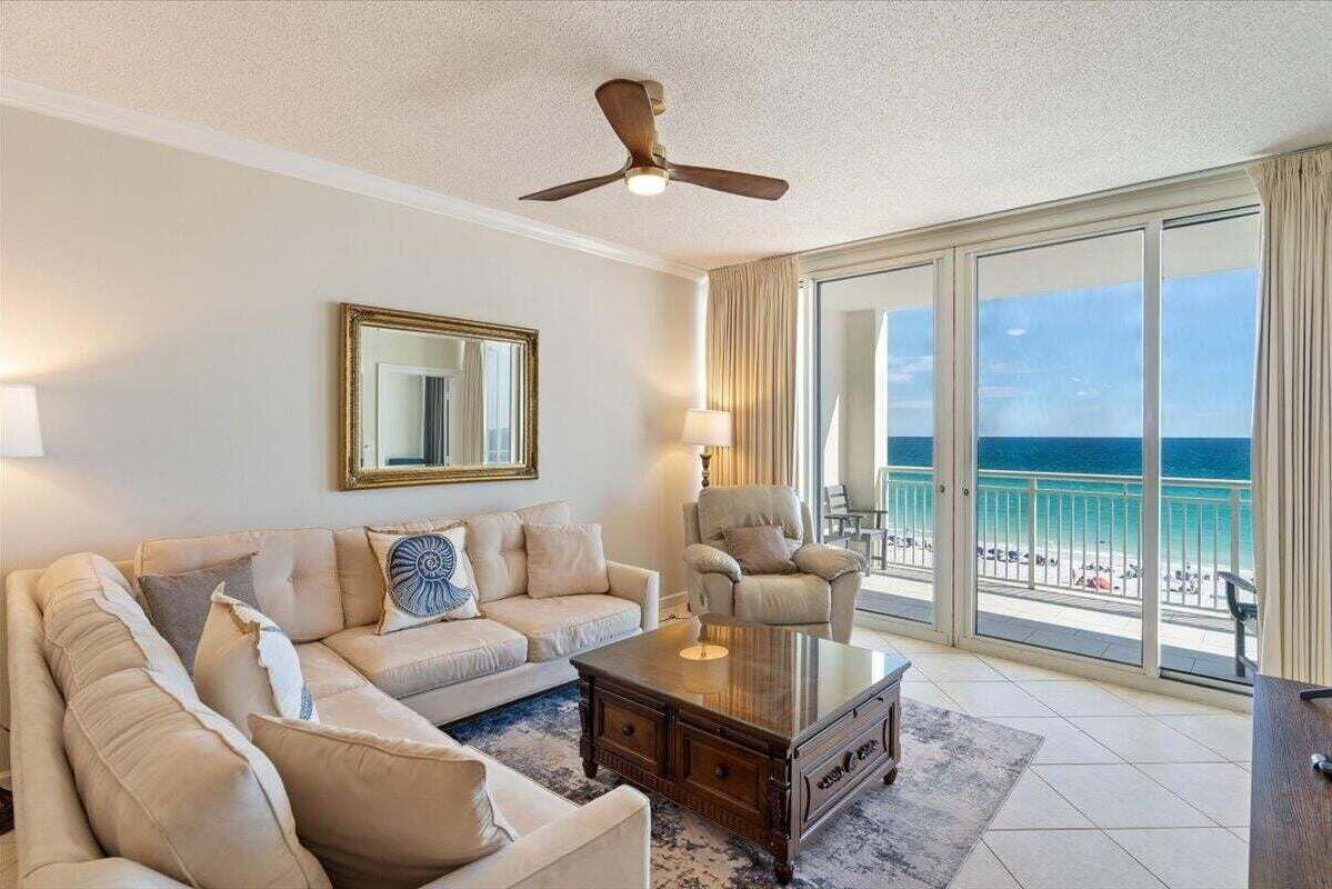 A living room with a sofa, armchair, and coffee table, a ceiling fan, a large mirror, and a view of the ocean through large sliding doors.
