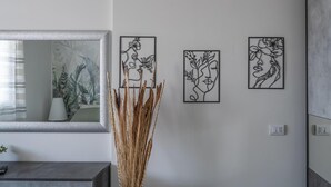 Moderni sisustus, jossa seinällä on kolme kehystettyä taideteosta, korkea koristeellinen maljakko ja peili, johon heijastuu kasvi.