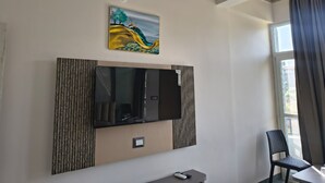 Et hotelværelse med et stort fladskærms-tv på væggen, et lille bord med en stol og et vindue med gardiner.