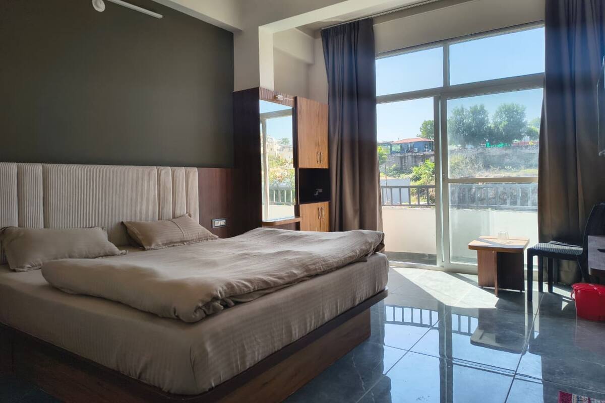 A modern hotel room with a large bed, a wooden wardrobe, a small table, and a balcony with a view.