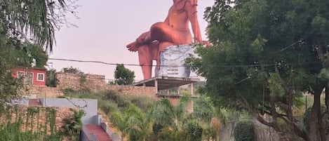 Una estatua roja de gran tamaño que representa una figura sentada en un parque, con césped, árboles y una estructura de piedra al fondo.