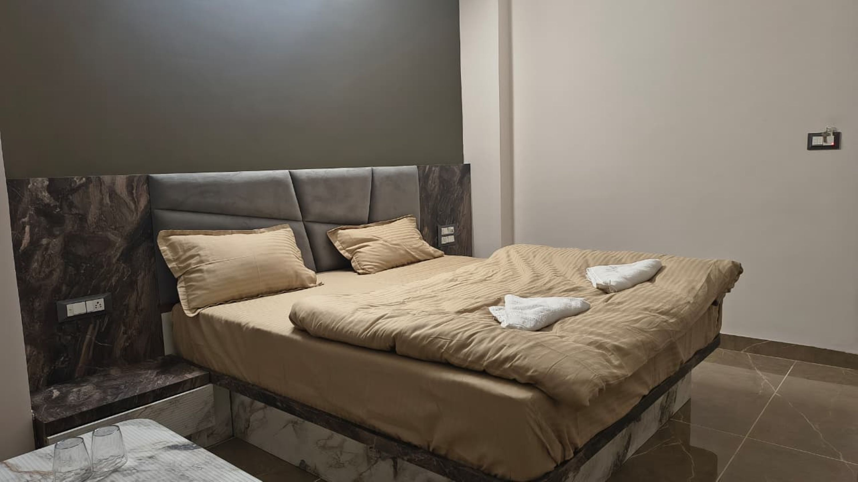 A neatly made bed with a headboard, a bedside table with glasses, and a marble-like floor.