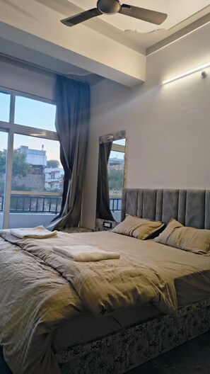 A bedroom with a large bed, a ceiling fan, and a view of the outside through a window. - Shree Vallabh Resort (Nathdwara)