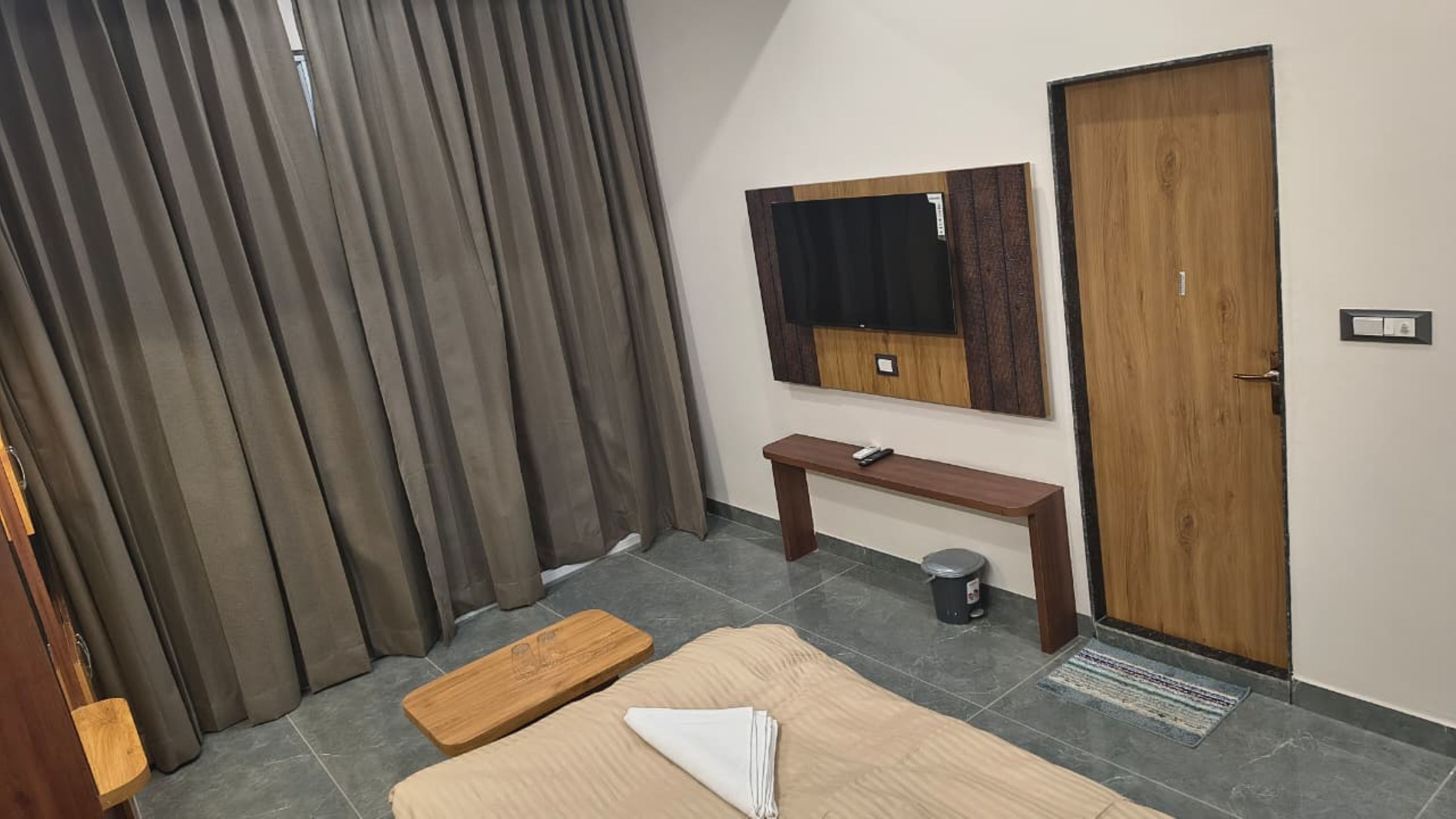 A hotel room with a large window, a wooden bench, a small table, a TV mounted on the wall, and a door.